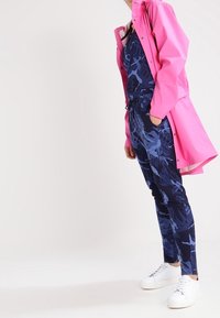 Cappotto impermeabile rosa brillante sopra una tuta a fantasia blu e pantaloni blu scuro. Sneakers bianche completano il look.