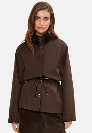 Frau trägt eine dunkelbraune Langarmjacke mit einem hohen glänzenden schwarzen Kragen und einem dünnen schwarzen Gürtel, der in der Taille gebunden ist.