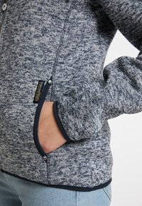 Maglione in pile grigio-blu con zip e tasca laterale, caratterizzato da rifiniture blu scuro sui polsini e sull'orlo, e un'etichetta del marchio vicino alla tasca.