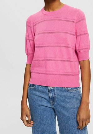 Person trägt einen rosa kurzärmeligen Strickpullover mit horizontalen Lochstreifen und blaue hoch taillierte Jeans.