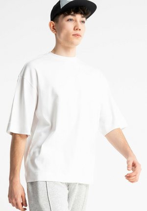 Młody mężczyzna ubrany w biały oversize t-shirt, szare spodnie i czarno-białą czapkę, patrzący przed siebie z neutralnym wyrazem twarzy.