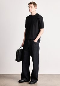 Fekete pamut rövid ujjú póló kis logóval, széles szárú sötét farmernadrággal párosítva. A modell egy fekete merev táskát tart.