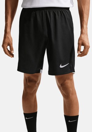 Mann trägt schwarze Nike-Sportshorts und schwarze Nike-Socken, steht mit entspannten Armen neben dem Körper vor einem weißen Hintergrund.