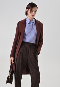 Donna vestita con un cappotto marrone, camicetta azzurro chiaro con fiocco, pantaloni scuri con dettagli brillanti, che tiene una borsa scura su uno sfondo neutro.