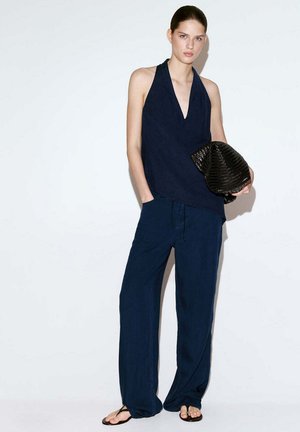 Mujer con un top halter azul marino y pantalones de pierna ancha, sosteniendo un bolso negro tejido, de pie frente a un fondo blanco liso.