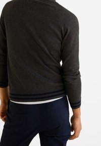 Homme portant un pull gris foncé avec des poignets et un ourlet côtelés, associé à un pantalon bleu marine, debout devant un fond blanc.