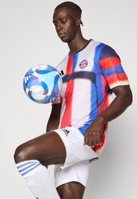 Φανέλα της Μπάγερν Μονάχου σε ζωντανό κόκκινο, μπλε και λευκό σχέδιο· κοντά μανίκια· λευκό σορτς που ταιριάζει με μαύρες ρίγες Adidas· ποδοσφαιρική μπάλα σε εστίαση.