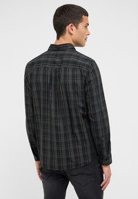 Ein Mann, der von hinten aussieht, trägt ein dunkelgrünes und schwarzes karierte Langarmhemd mit orangenen Linien und dunkelgrauen Jeans vor einem schlichten Hintergrund.