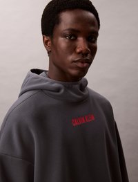Felpa grigia con cappuccio e finitura testurizzata, caratterizzata da un logo rosso "CALVIN KLEIN" sulla parte anteriore, progettata con una vestibilità rilassata e un ampio scollo.