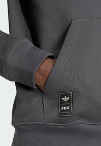 Χέρι στην μπροστινή τσέπη μιας σκουρόχρωμης γκρι κουκούλας με ετικέτα Adidas FIFA Classics κοντά στο τελείωμα.