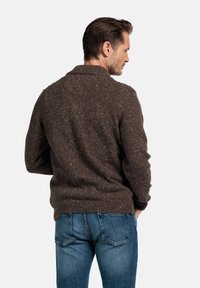 Bruine, textured cardigan met een gespikkeld patroon, ribbelmanchetten en zoom. Draagt over blauwe spijkerbroeken, gezien van achteren.