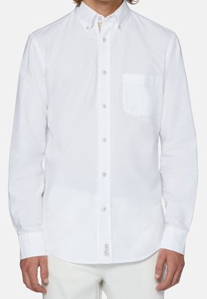 Man wearing a long-sleeve white button-up shirt with a chest pocket and white pants, standing against a plain background.