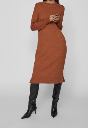 Femme portant une longue robe en maille couleur rouille avec une fente latérale et des bottes en cuir noires à talons montant jusqu'aux genoux, se tenant devant un fond uni.
