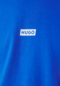 Sinine puuvillane t-särk, millel on valge ristkülikukujuline logo vasakul küljel tekstiga "HUGO". Sile tekstuur, lihtne disain.