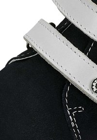 Scarpa in suede nera con cinturini in pelle bianca, cuciture a contrasto e un dettaglio circolare lucido. Primo piano degli elementi di design della scarpa.