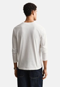 Homme aux cheveux courts et bouclés portant une chemise blanche unie à manches longues et un jean foncé, vu de dos sur un fond blanc.