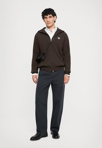 Brown zip-up jacket with logo, layered over a white shirt, paired with dark pants and black shoes. Small black crossbody bag.
