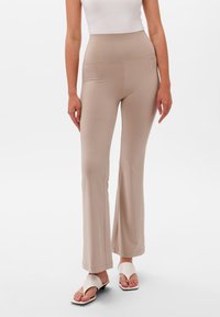 Beżowe spodnie z szerokimi nogawkami i wysokim stanem, wykonane z gładkiego, elastycznego materiału. Noszone z białymi płaskimi sandałami o minimalistycznym designie.