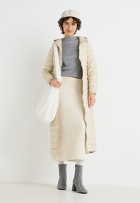 Abrigo acolchado beige, suéter de canalé gris, falda de punto crema, botas de tacón grises y un bolso blanco texturizado. Sombrero en color beige a juego.