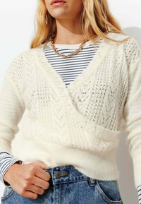 Cardigan blanc en tricot avec un col en V et un motif en chevrons, superposé à un top rayé bleu et blanc. Collier en chaîne dorée en accent.