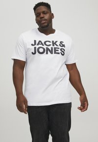 Bijela kratka majica s kratkim rukavima s velikim crnim natpisom "JACK & JONES" otisnutim sprijeda. Mekana tkanina, okrugli izrez, ležeran kroj. Tamni hlače su vidljive.
