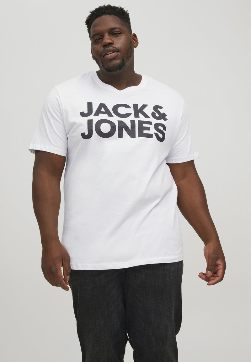 Bijela kratka majica s kratkim rukavima s velikim crnim natpisom "JACK & JONES" otisnutim sprijeda. Mekana tkanina, okrugli izrez, ležeran kroj. Tamni hlače su vidljive.