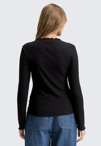 Zwarte langemouwen top met ribbeltextuur, gegolfde halslijn en zoom. De achterkant laat een getailleerd ontwerp zien in combinatie met high-waisted denim.
