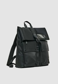 Zaino nero in nylon liscio con chiusura a patta, due tasche anteriori e spallacci regolabili. Presenta una finestra trasparente per il badge identificativo sulla parte anteriore.