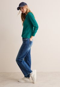 Tiefblaue Langarmbluse kombiniert mit dunkelblauen Straight-Leg-Jeans. Weiße Sneakers und eine marineblaue Kappe runden das Outfit ab.