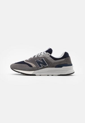 Grå og navy New Balance sneakers med hvide snørebånd, ruskind og mesh overdel, polstret sål og det mærkede "N" logo på siden.