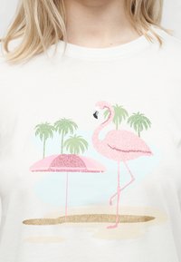 Ροζ φλαμίνγκο με γκλίτερ, που στέκεται στο ένα πόδι δίπλα σε μια ροζ ομπρέλα σε μια παραλία με φοίνικες στο φόντο σε λευκό μπλουζάκι.