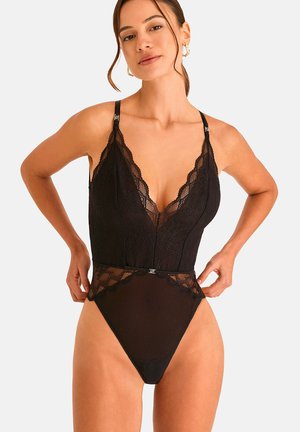 Femme portant un body en dentelle noire avec des panneaux en mesh transparent et des bretelles réglables, debout devant un fond clair uni.