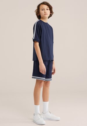 Băiat stând în picioare, purtând un tricou sport bleumarin cu dungi albe pe mâneci, pantaloni scurți asortați cu un bord alb, șosete albe și adidași albi.