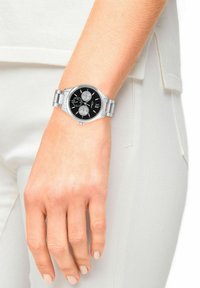 Silver metal bracelet watch with a black dial, three sub-dials, and silver accents. Displaying a minimalist design, set against light-colored fabric.