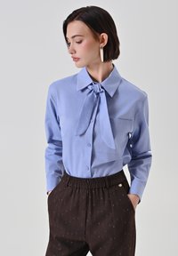 Blusa azzurro chiaro con colletto a fiocco, tasca frontale e chiusura a bottoni; pantaloni neri con motivo a righe e vita elasticizzata.