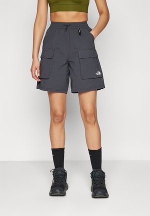 Persona vistiendo pantalones cortos cargo gris oscuro con grandes bolsillos, calcetas negras y zapatos de senderismo negros, de pie con una mano en un bolsillo.