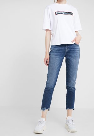 Mujer con camiseta blanca de gran tamaño con el logo de "Opening Ceremony", vaqueros azules recortados con dobladillos deshilachados y zapatillas blancas gruesas.