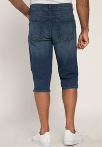 Pantalons capri en denim, bleu foncé, mi-mollet avec deux poches arrière, coutures à double surpiqûre et ourlet retroussé. Portés avec des baskets blanches.