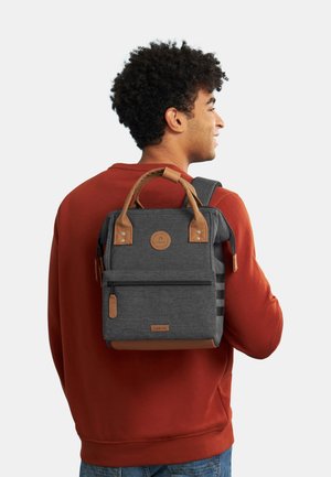 Sac à dos en tissu gris avec des accents en cuir brun, forme rectangulaire, poignée supérieure, sangle réglable, poche zippée à l'avant, patch logo visible.
