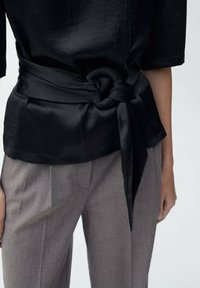 Haut en satin noir avec une ceinture nouée à la taille, associé à un pantalon gris clair sur mesure avec des plis à l'avant. Texture lisse et design minimaliste.