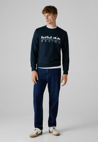 Sudadera navy con el logo de "Red Bull Racing", cuello redondo, puños acanalados, combinada con unos vaqueros azul oscuro y zapatillas blancas con colores de acento.