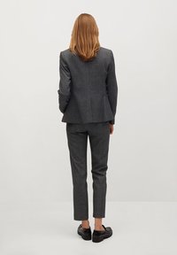 Costume long et ajusté en tissu texturé gris foncé, comprenant une veste et un pantalon fuselé. Des chaussures noires complètent l'ensemble, vu de l'arrière.