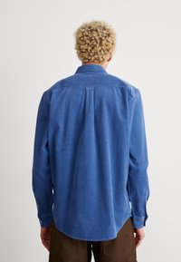 Camisa azul de pana con cuello abotonado, mangas largas y dobladillo curvado; presenta textura de costillas verticales y puños ajustables.