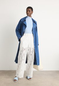 Trench coat in denim, dolcevita azzurro chiaro, pantaloni a gamba larga bianchi con occhietti, e sandali azzurri chiari, con una borsa crema in mano.