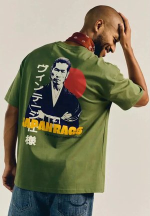 Man lacht met hand op zijn hoofd, draagt een groen T-shirt met een afbeelding van een strenge samoerai met een rode cirkel en de tekst "JAPAN RAGS" op de achterkant.