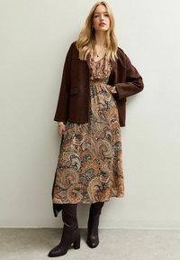 Bruine suède jack over een gemêleerde maxi-jurk met warme tinten en ingewikkelde paisleypatronen. Kniehoge donkerbruine laarzen maken de outfit compleet.