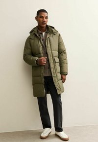 Abrigo acolchado en verde oliva, diseño de acolchado, hasta la rodilla, con cierre de cremallera y botones, combinado con una camisa a cuadros, vaqueros negros y zapatillas blancas.