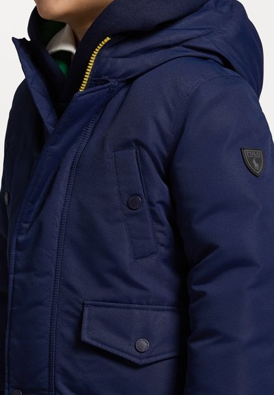 Ναυτικό μπλε μπουφάν με κουκούλα, κλείσιμο με φερμουάρ και κίτρινη λεπτομέρεια, καθώς και δύο μπροστινές τσέπες, διακοσμημένο με ένα μαύρο λογότυπο.