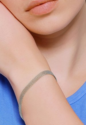 Primo piano del polso di una persona che indossa un braccialetto a maglie d’argento, appoggiato vicino al mento, su uno sfondo di camicia blu.