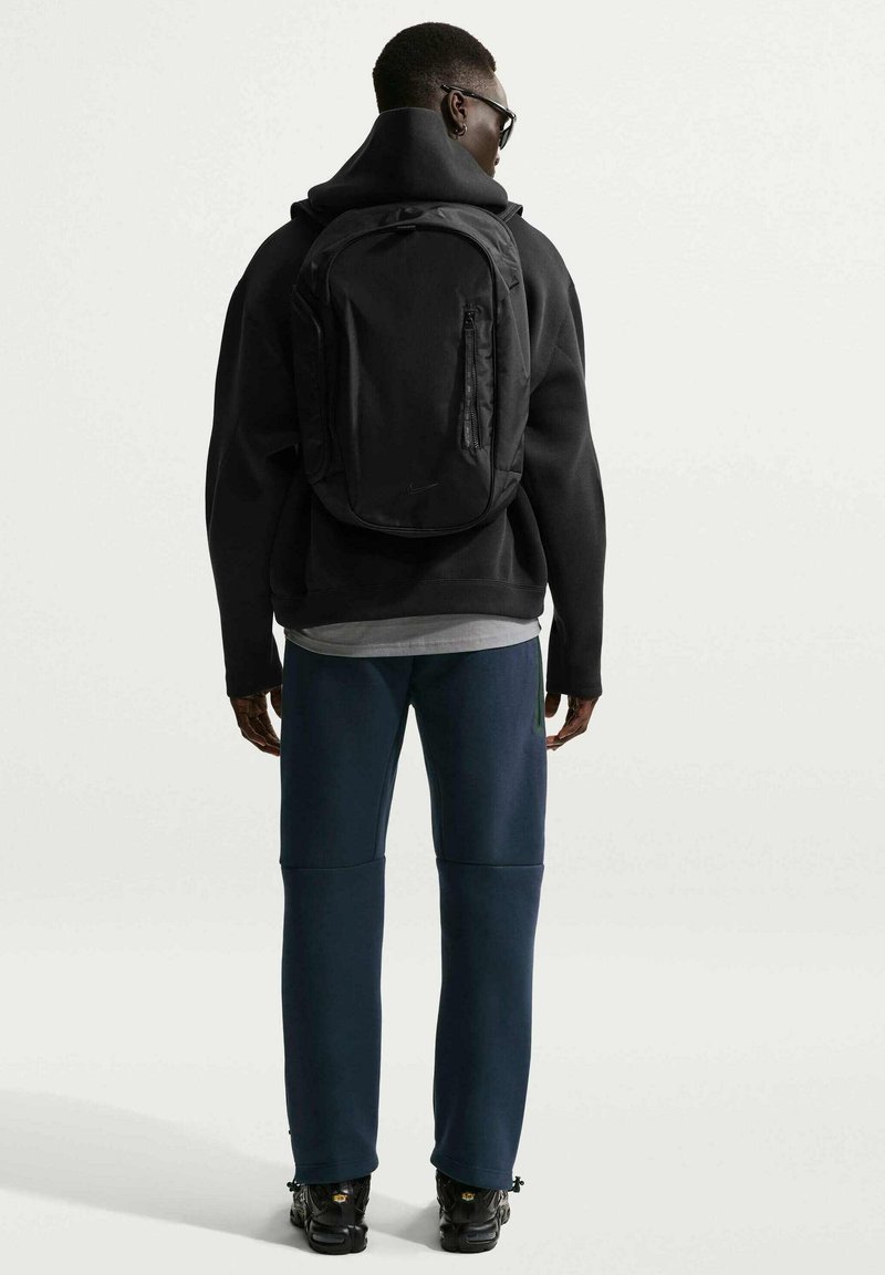 Mochila negra de diseño elegante, con bolsillo frontal con cremallera y correas acolchadas, llevada por una persona con sudadera negra con capucha y pantalones deportivos azules.